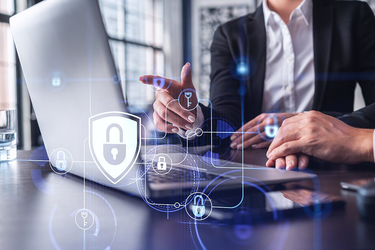 A closeup view of a laptop has cybersecurity symbols superimposed over it. A businesswoman is behind, slightly out of focus, as she points to the laptop.